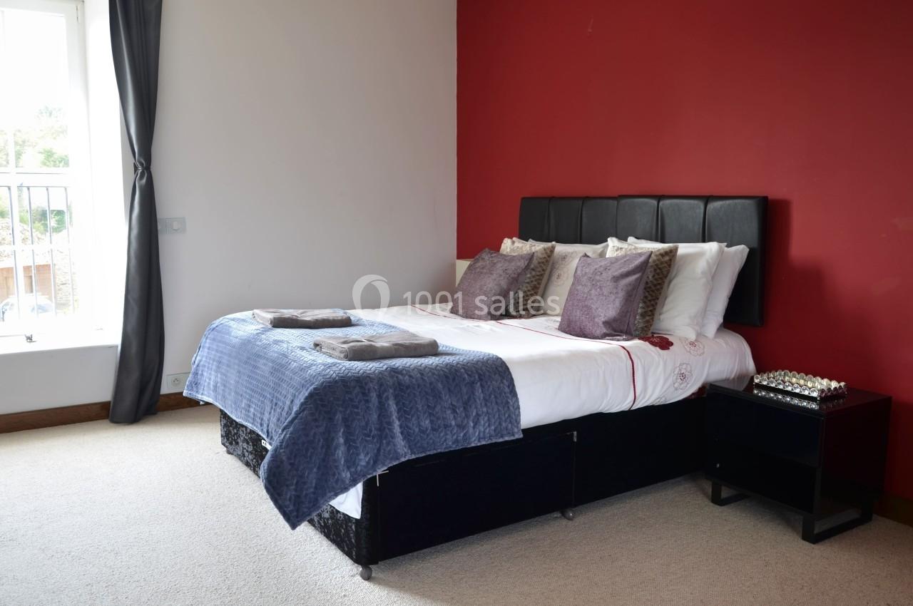 Chambre avec un lit double, draps blancs, coussins décoratifs, mur rouge, table de chevet et fenêtre avec rideaux gris.