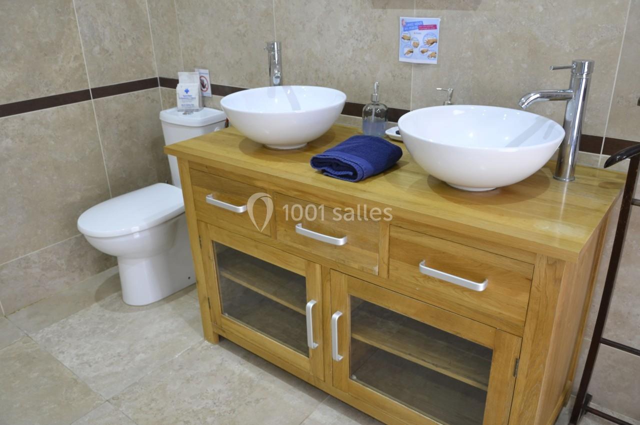 Meuble de salle de bain en bois avec deux vasques blanches, un miroir absent, et une toilette en arrière-plan.