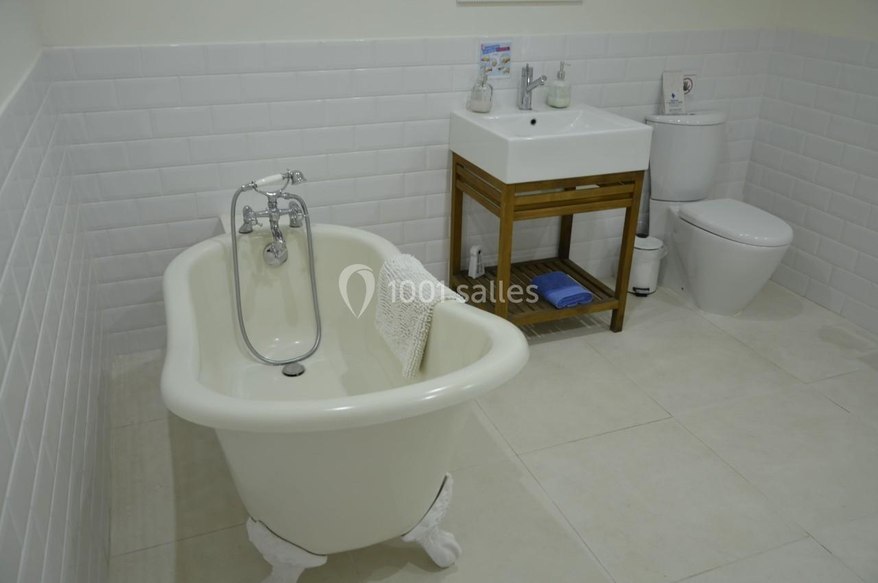 Salle de bain avec baignoire sur pieds, lavabo avec meuble en bois, toilettes et carrelage blanc.