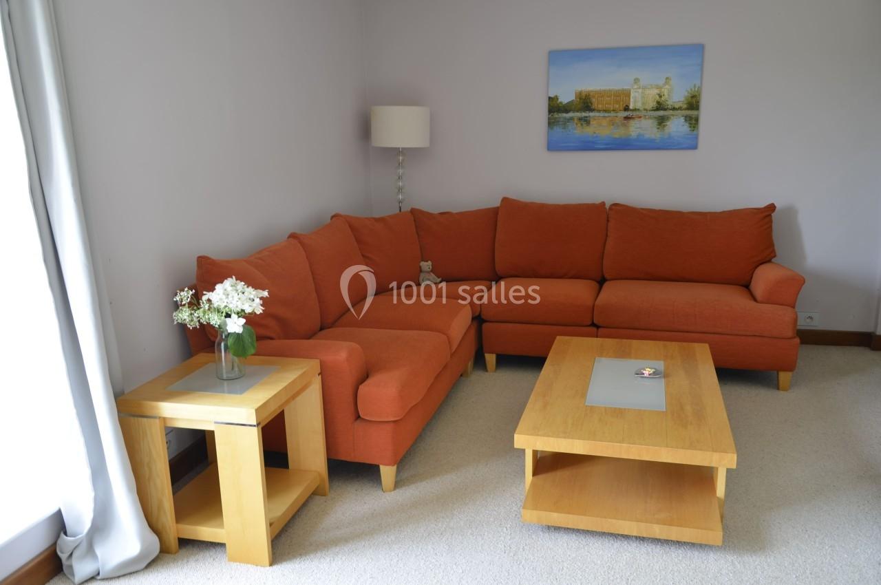 Salon avec canapé d'angle orange, table basse en bois, lampe sur pied et tableau mural représentant un paysage.