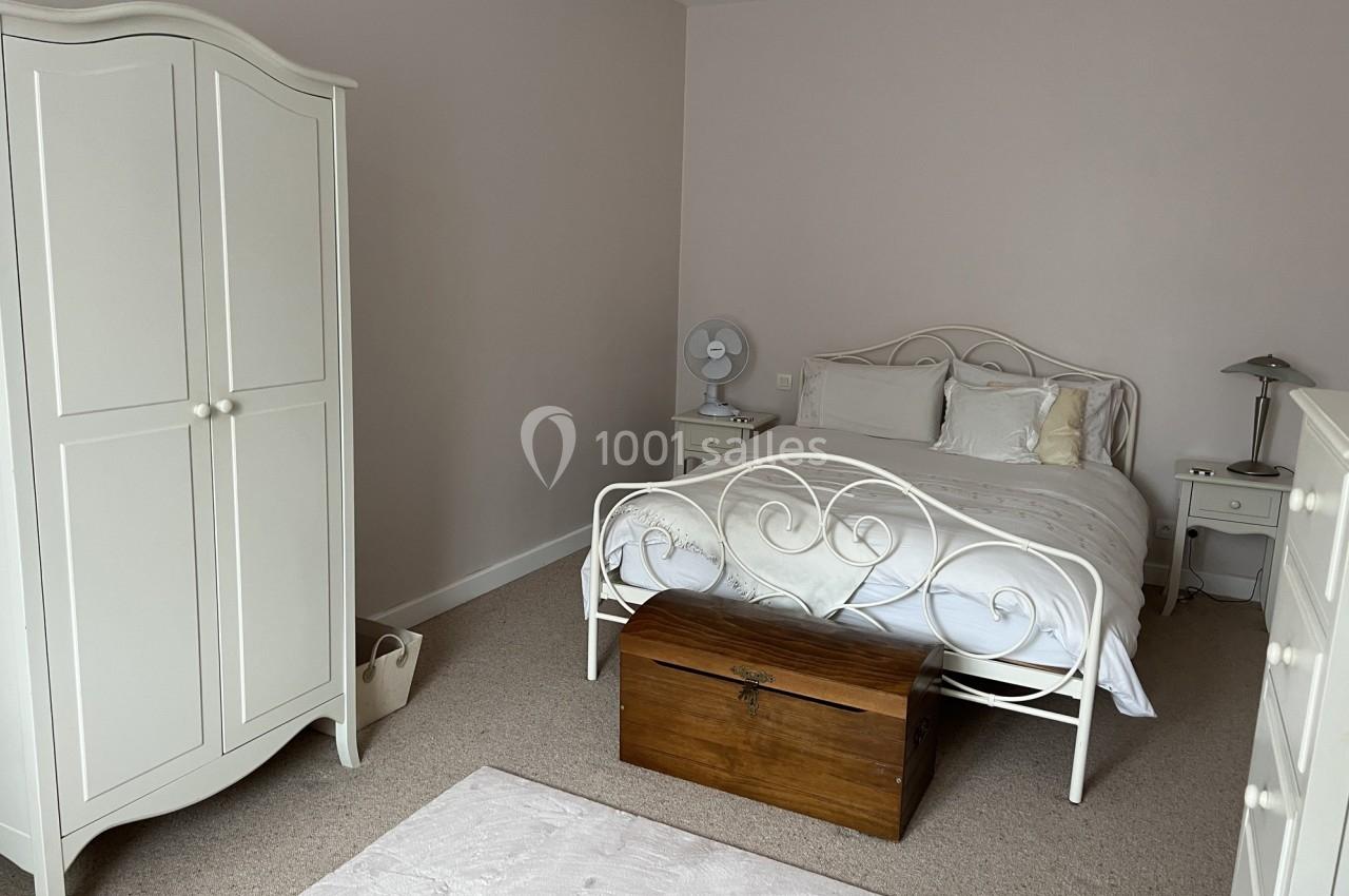 Chambre lumineuse avec lit double en métal blanc, armoire assortie, coffre en bois et tapis clair sur moquette beige.