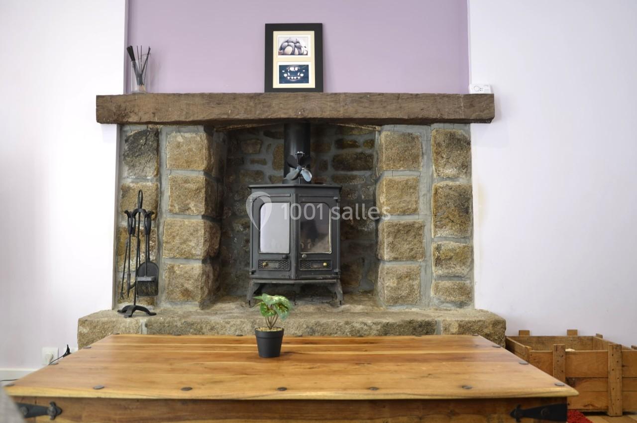 Cheminée en pierre avec un poêle à bois noir, une table en bois au premier plan et un mur violet en arrière-plan.