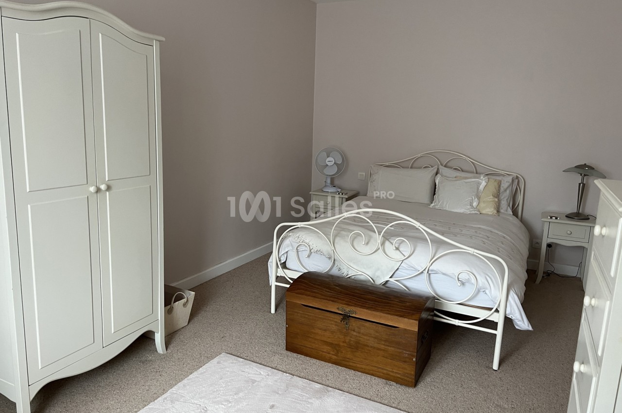 Chambre lumineuse avec lit double en métal blanc, armoire assortie, coffre en bois et tapis clair sur moquette beige.