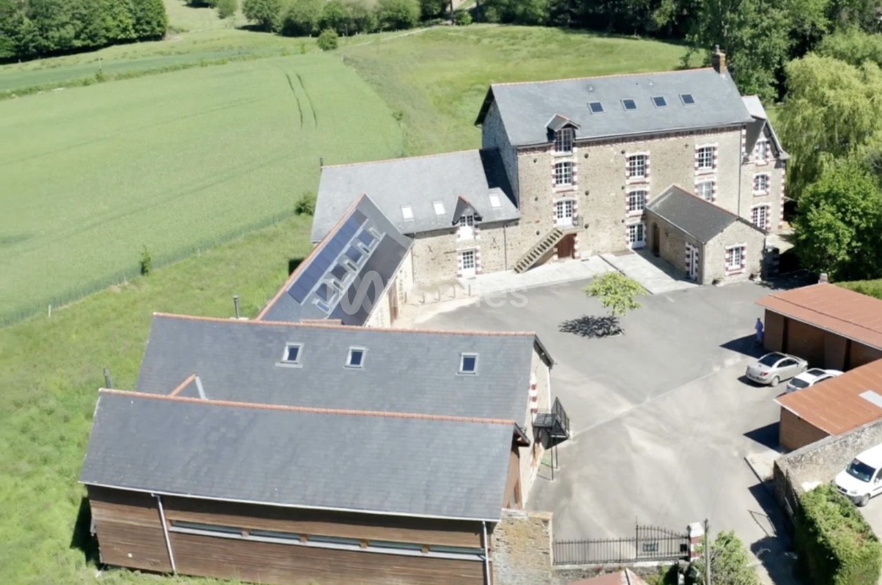 Vue aérienne d'un bâtiment en pierre avec dépendances modernes, entouré de champs et d'arbres.