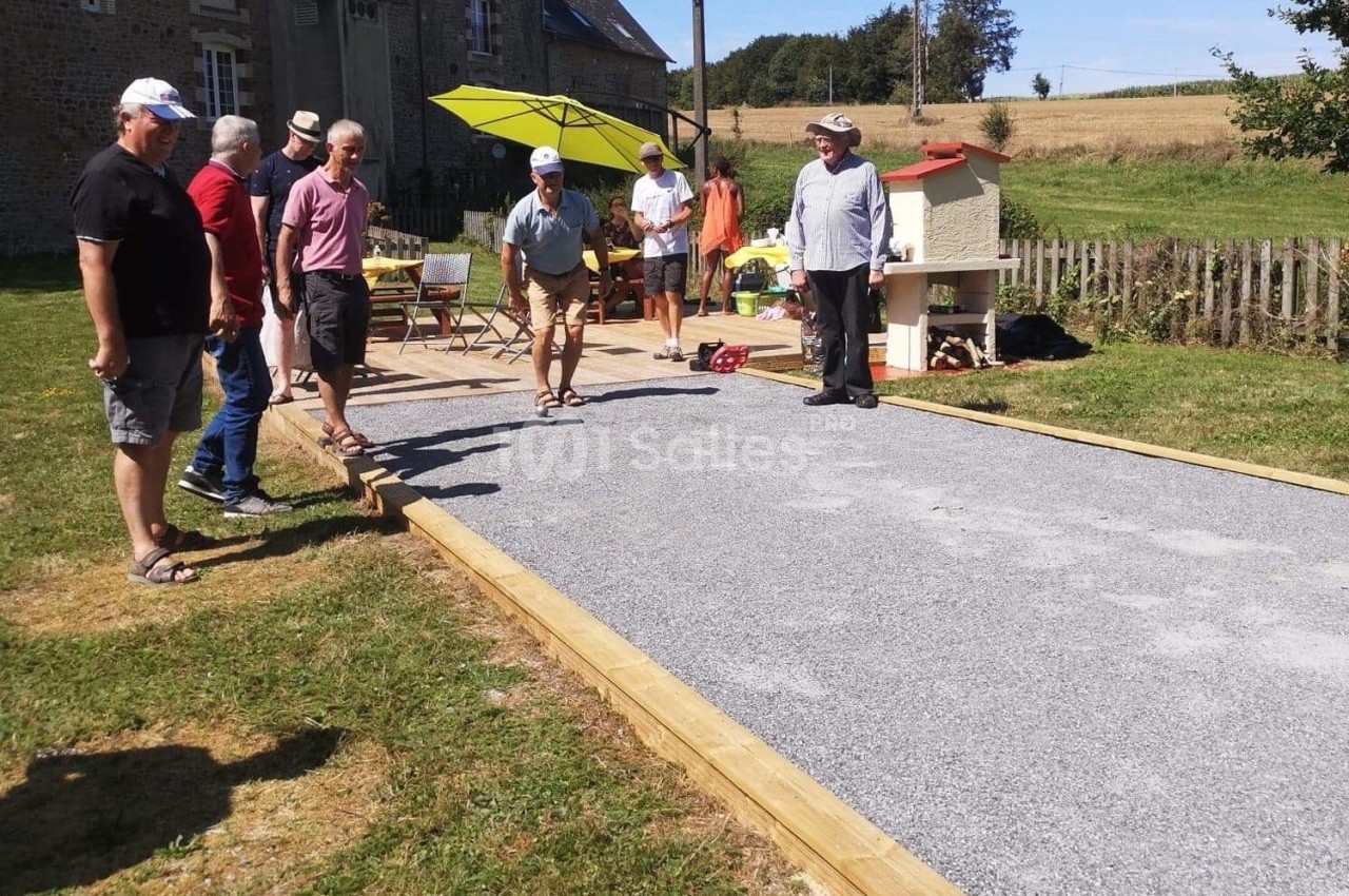 Des personnes jouent à la pétanque sur un terrain aménagé en plein air, entouré de verdure et de bâtiments ruraux.