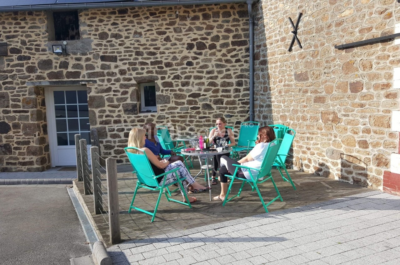Quatre personnes assises autour d'une table sur une terrasse en bois, devant une maison en pierre.