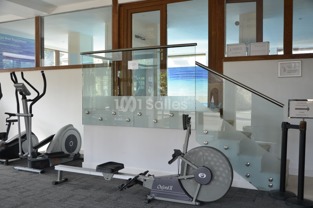 Salle de sport avec rameur, vélo d'exercice et escalier en verre dans un espace lumineux.