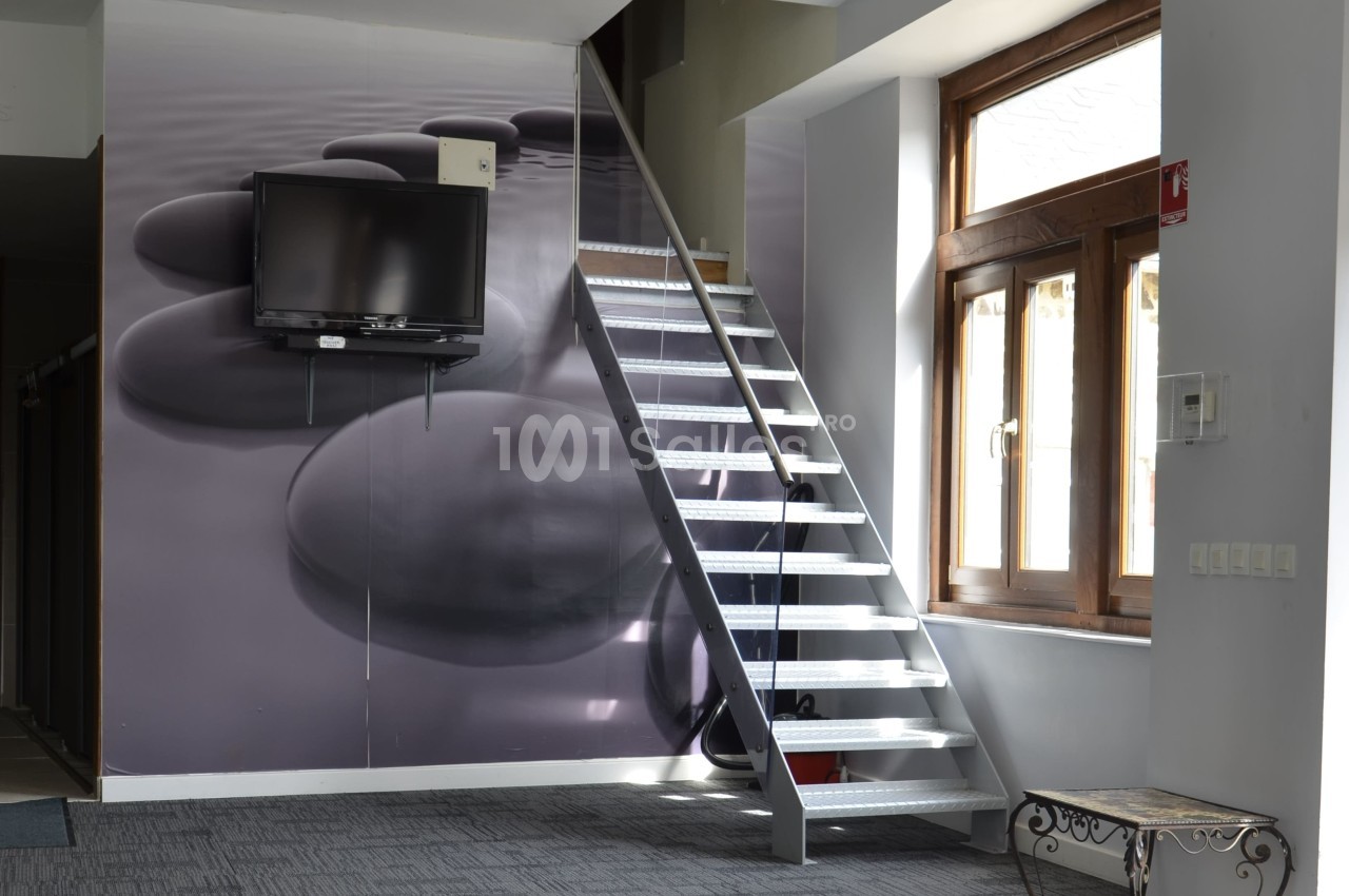 Escalier métallique intérieur près d'une fenêtre, avec un mur décoré de galets et une télévision fixée dessus.