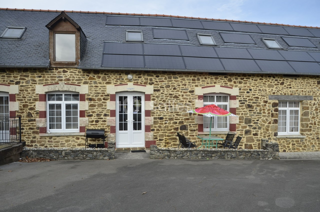 Façade en pierre d'une maison avec fenêtres blanches, toit en ardoise et panneaux solaires, table et parasol devant.