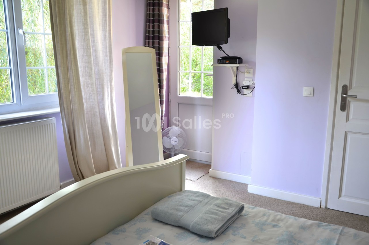 Chambre lumineuse avec lit, miroir, télévision murale, ventilateur au sol et fenêtres donnant sur un jardin.