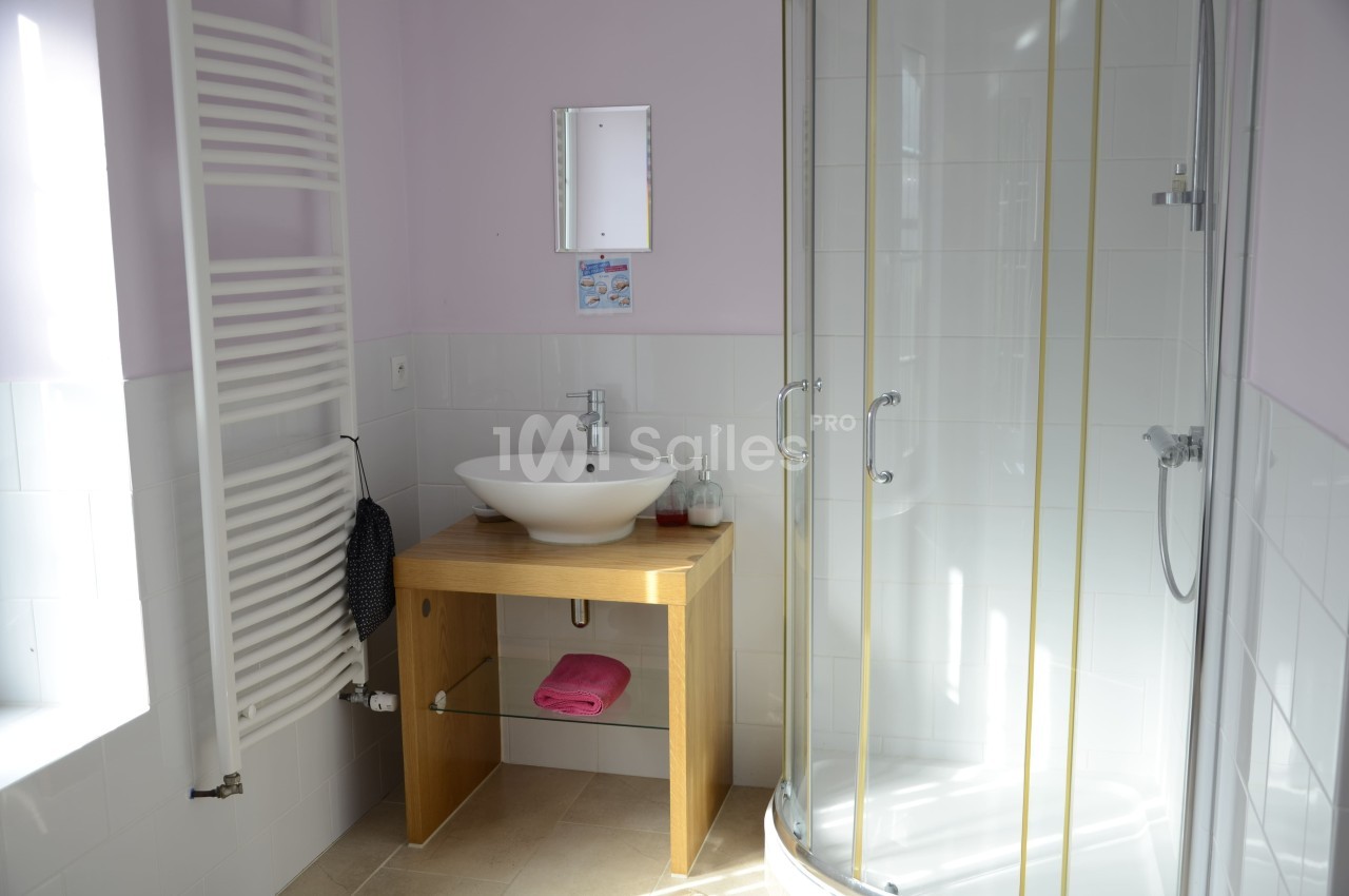 Salle de bain avec lavabo sur meuble en bois, douche vitrée, radiateur sèche-serviettes et mur peint en rose clair.