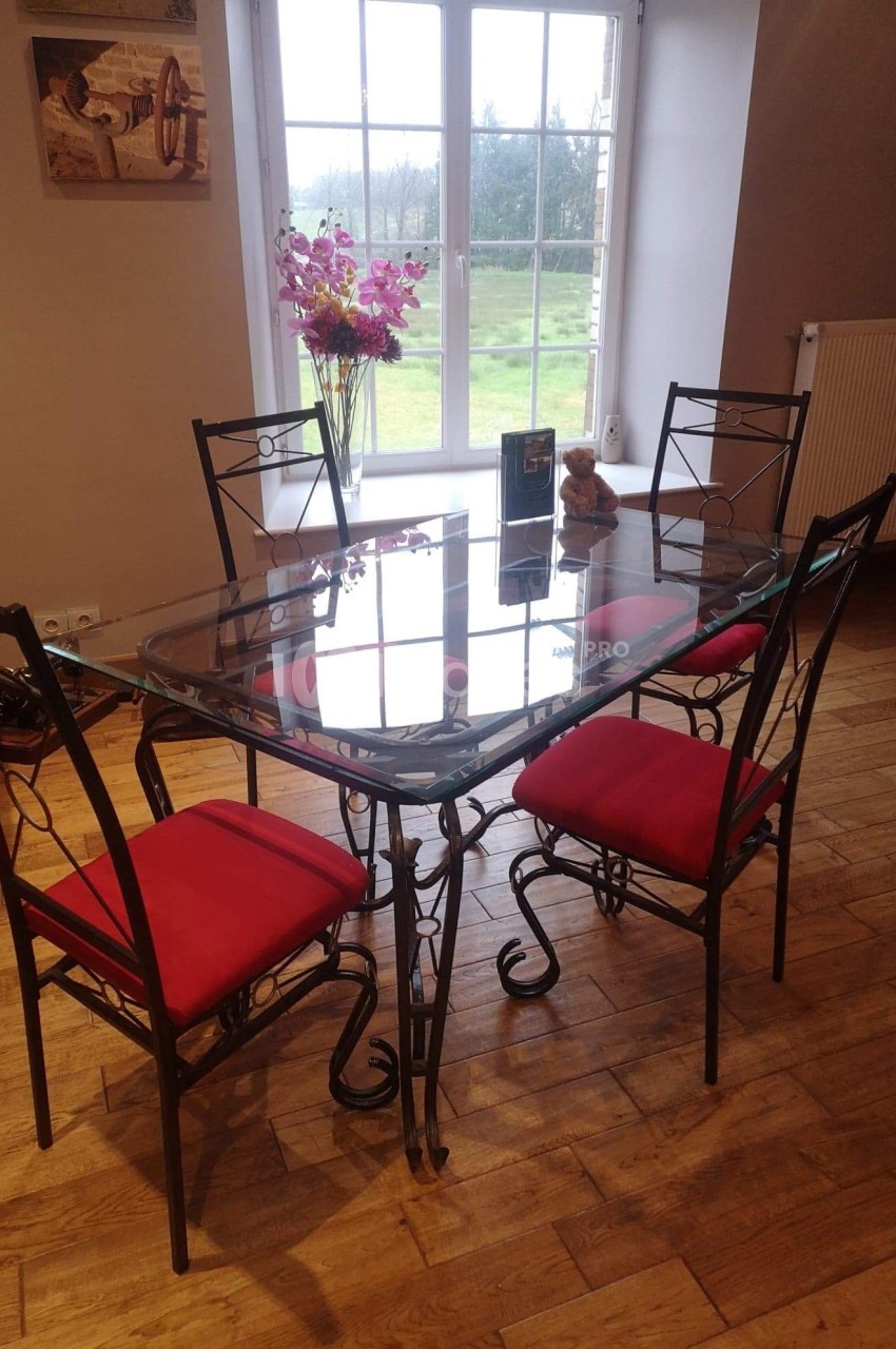Table en verre avec structure en métal noir, entourée de quatre chaises à coussins rouges, dans une pièce lumineuse.