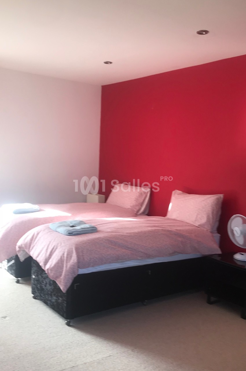 Deux lits simples avec draps roses dans une chambre lumineuse aux murs rouges et blancs, rideaux rouges et ventilateur sur…