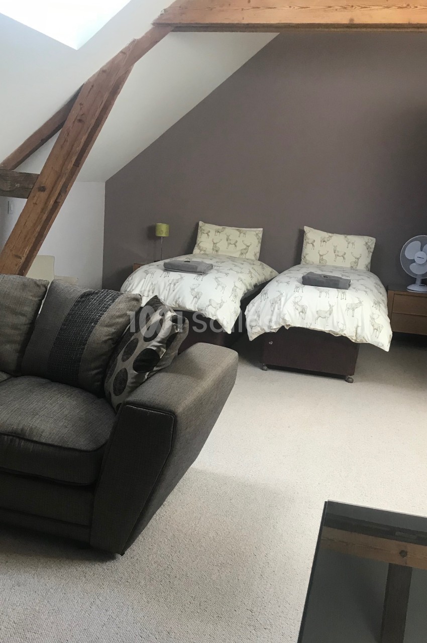 Chambre mansardée avec deux lits simples, canapé, table basse, ventilateur et poutres apparentes en bois.