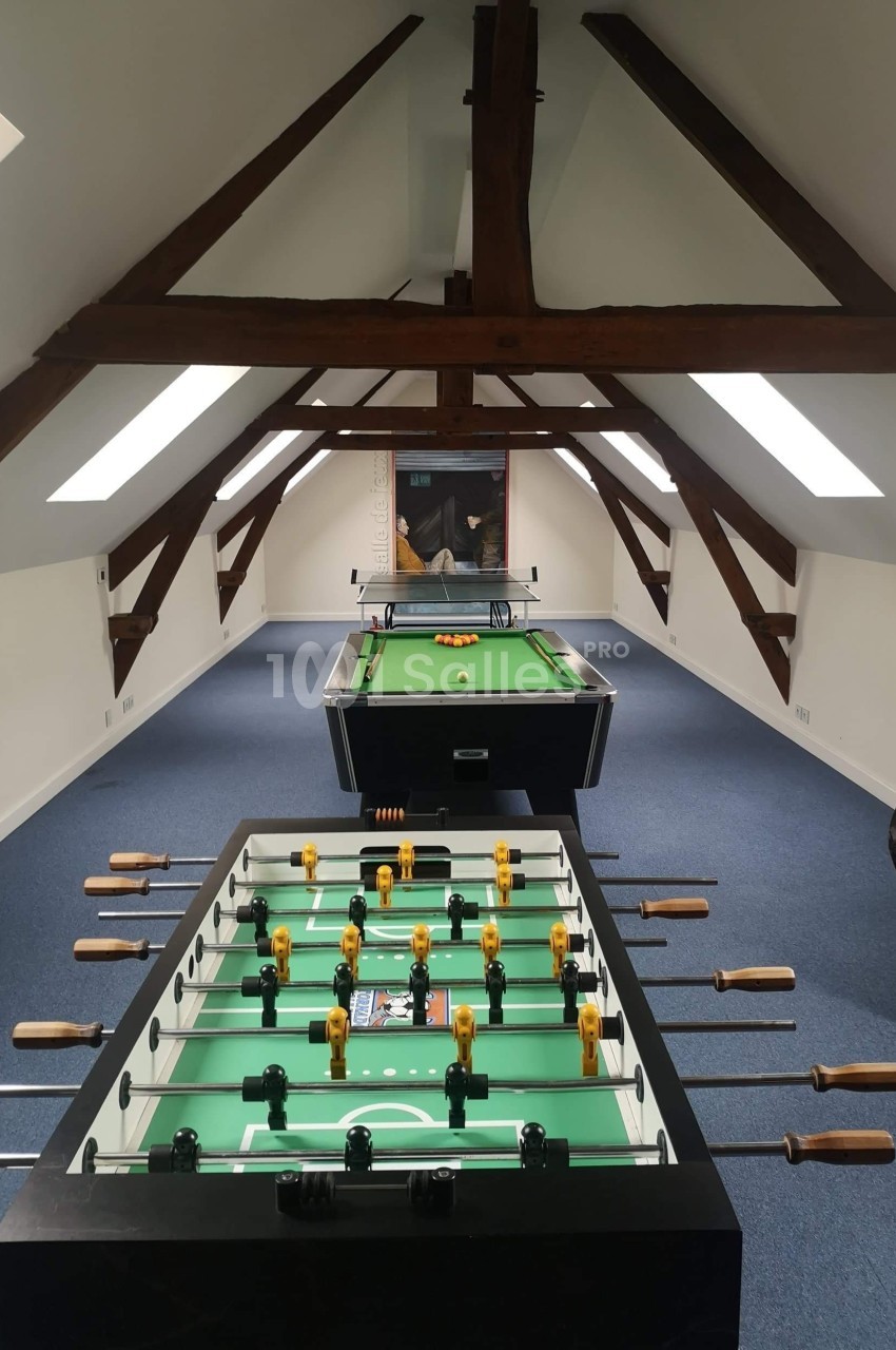 Salle mansardée avec baby-foot et billard alignés sur un sol en moquette bleue, éclairée par des fenêtres de toit.
