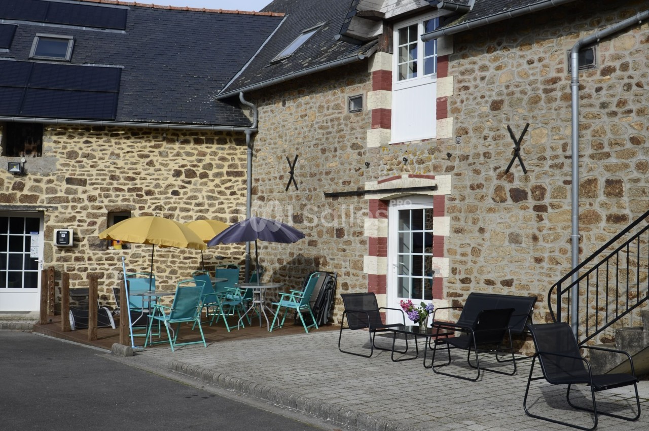 Terrasse avec tables, chaises colorées et parasols devant une maison en pierre avec volets blancs et rouges.