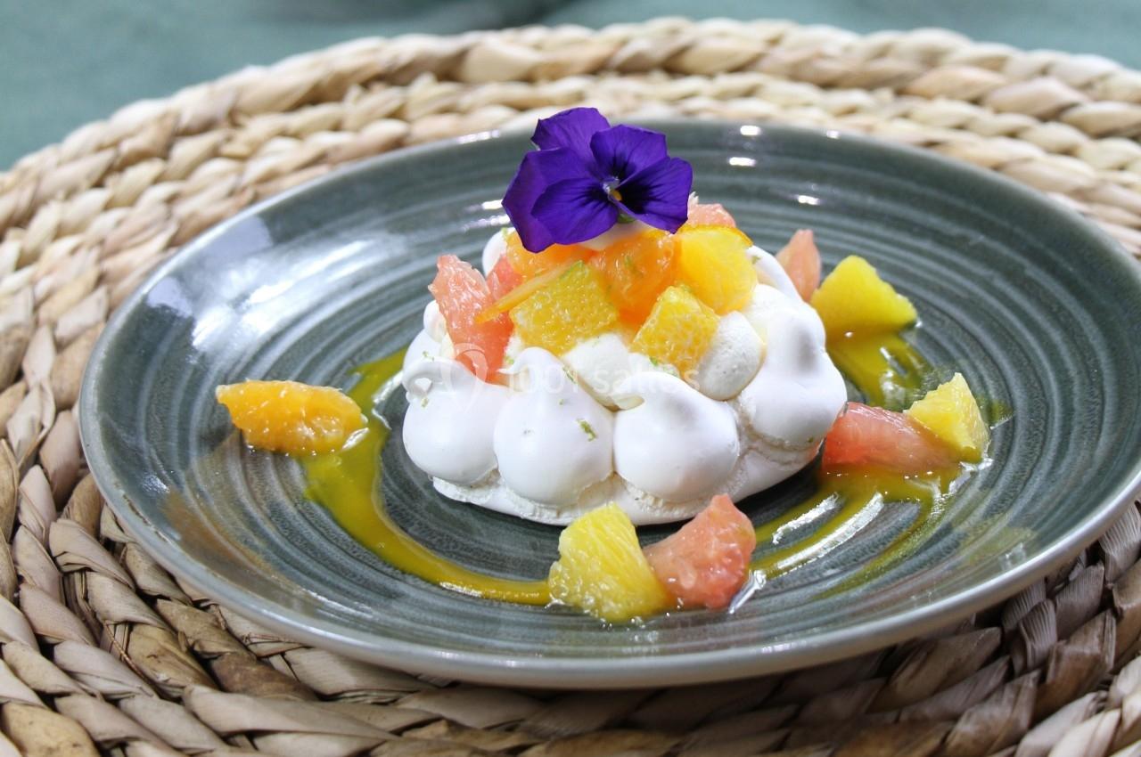 Dessert meringué garni de morceaux d'agrumes, nappé de coulis et décoré d'une fleur violette, sur une assiette grise.