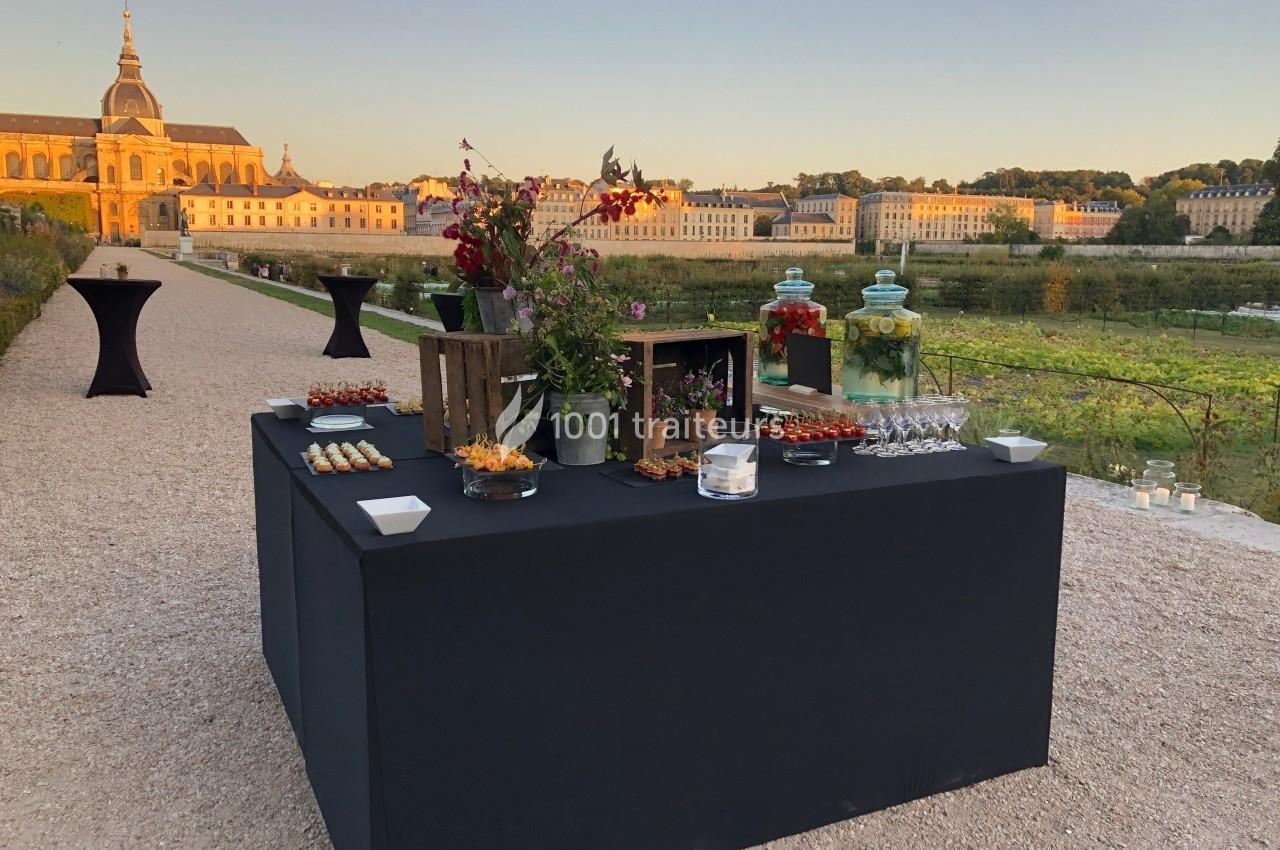 Table de buffet dressée en extérieur avec des amuse-bouches, entourée d'un paysage de château et de jardins au coucher du…