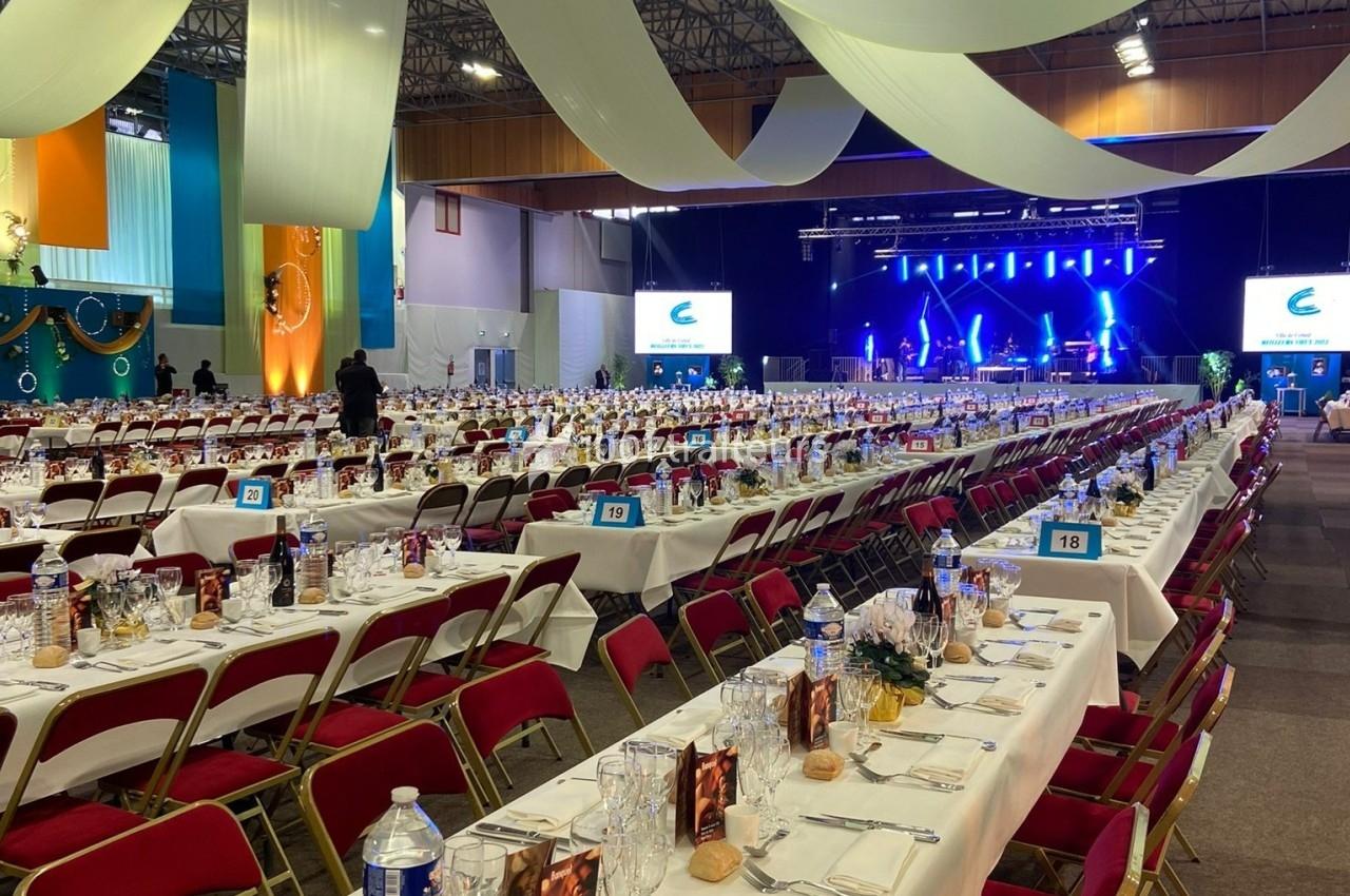 Salle de réception avec de longues tables dressées, chaises rouges et scène éclairée en arrière-plan.