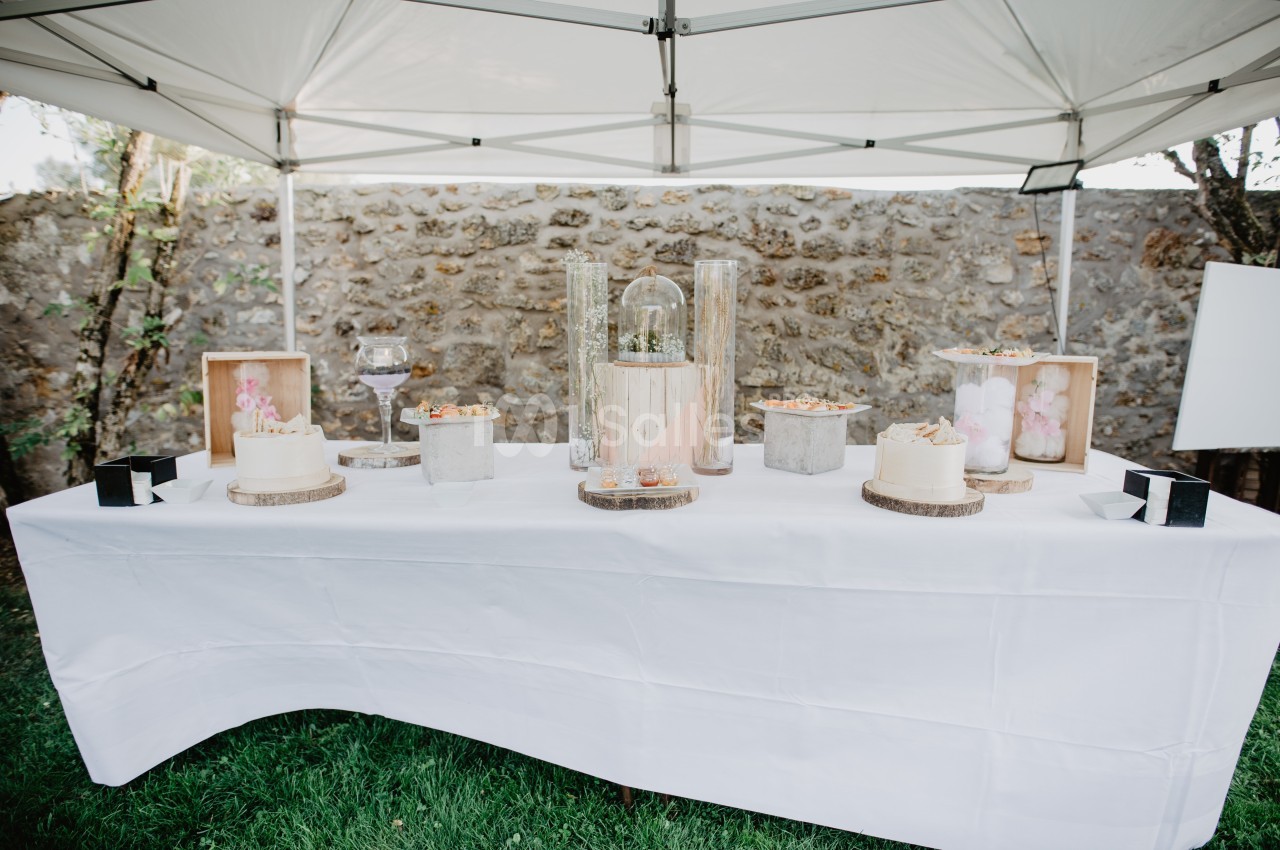 Table décorée sous une tente blanche avec des gâteaux, des fleurs et des éléments décoratifs sur fond de mur en pierre.
