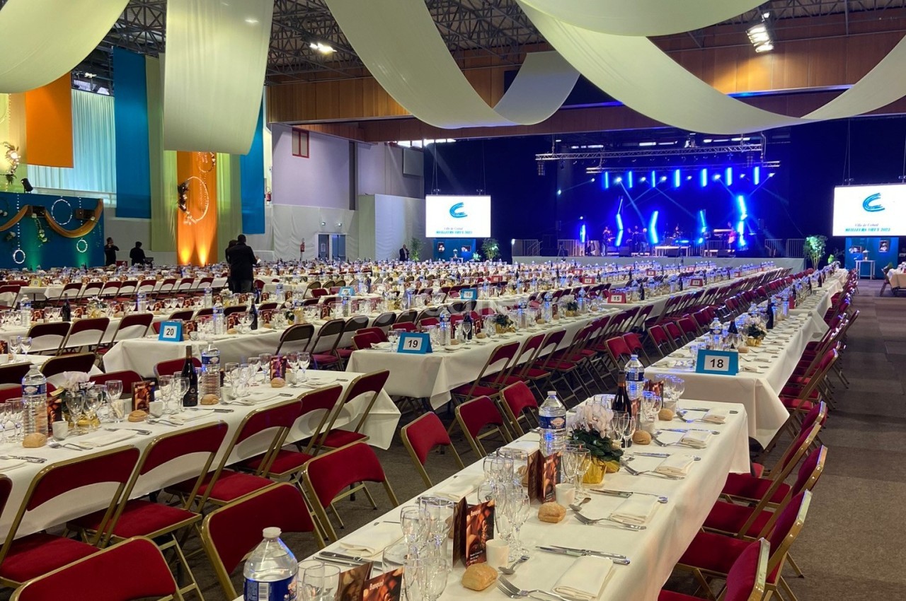 Salle de réception avec de longues tables dressées, des chaises rouges et une scène éclairée en arrière-plan.