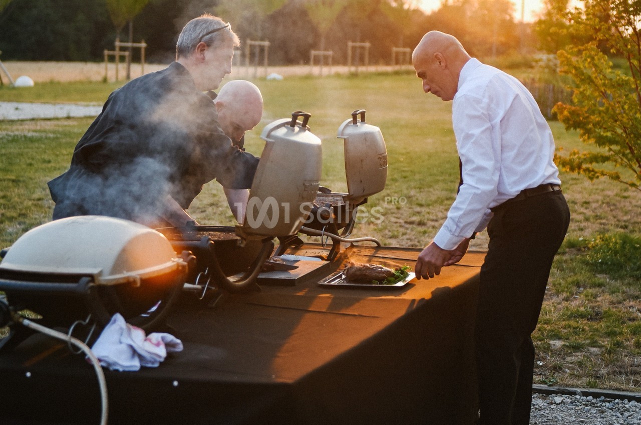 Trois hommes préparent des plats sur des barbecues en plein air, dans un cadre naturel au coucher du soleil.