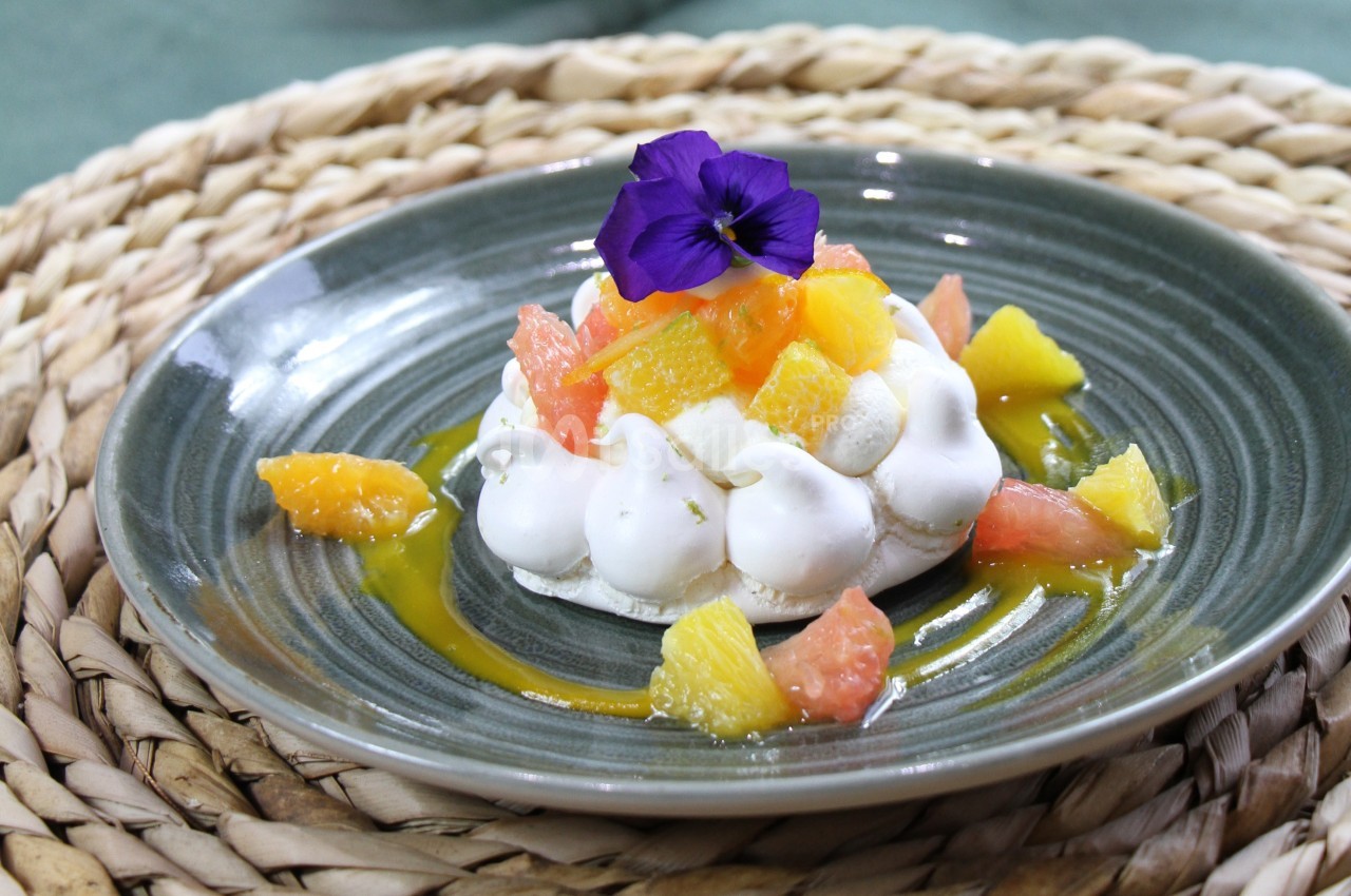 Dessert pavlova garni de morceaux d'agrumes colorés et décoré d'une fleur violette, présenté sur une assiette grise.