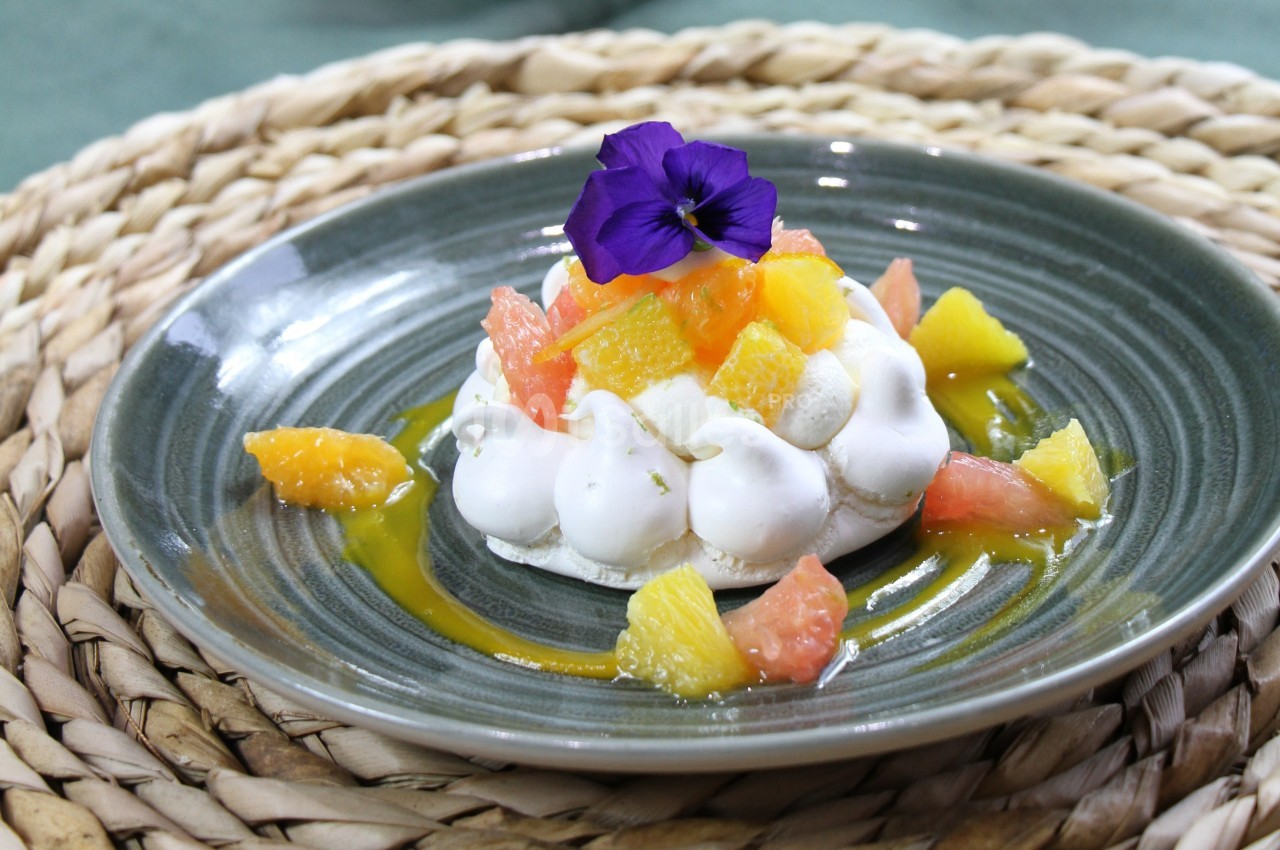 Dessert à base de meringue, agrumes variés et fleur comestible, présenté sur une assiette grise.