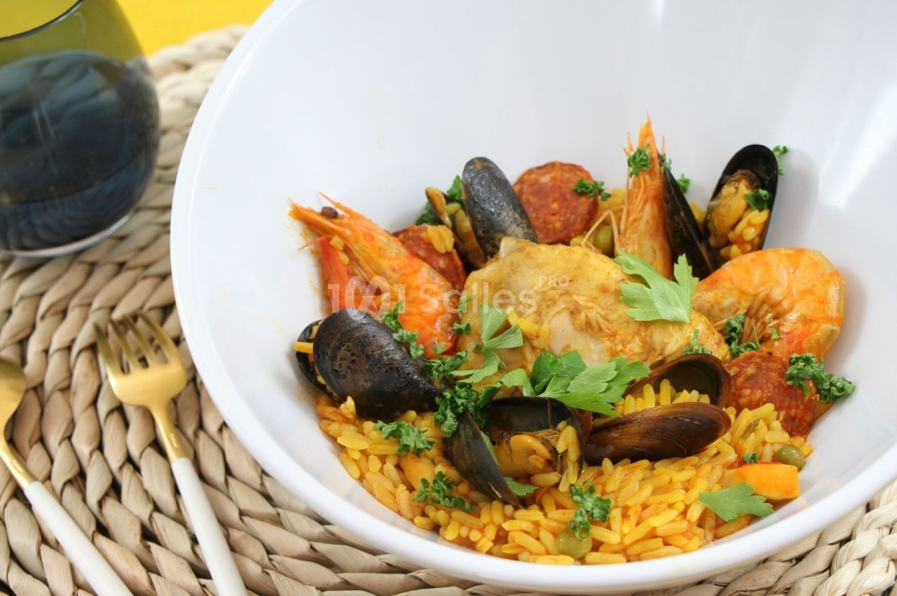 Plat de paella dans un bol blanc, garni de crevettes, moules, poulet, chorizo et persil, posé sur un dessous en osier.