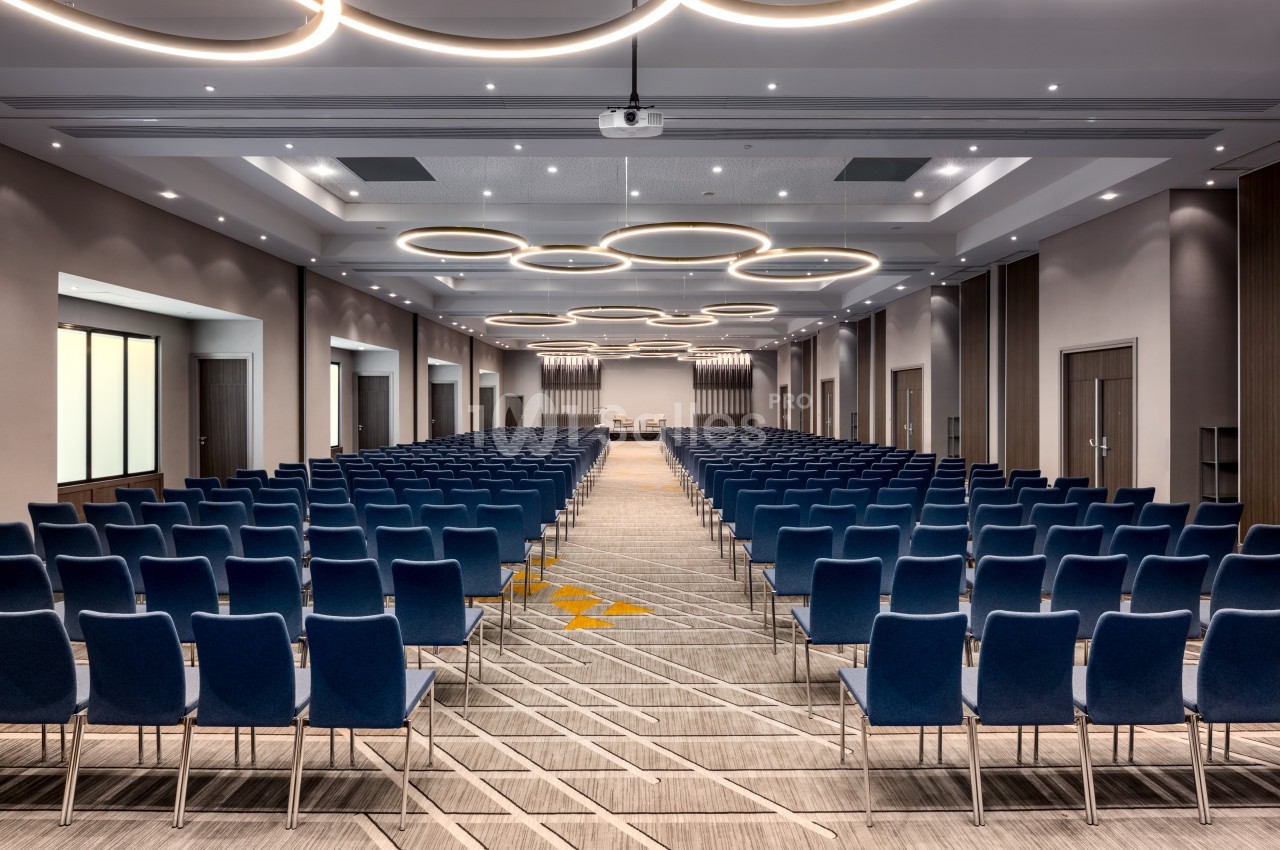 Salle de conférence moderne avec des rangées de chaises bleues alignées et des luminaires circulaires au plafond.