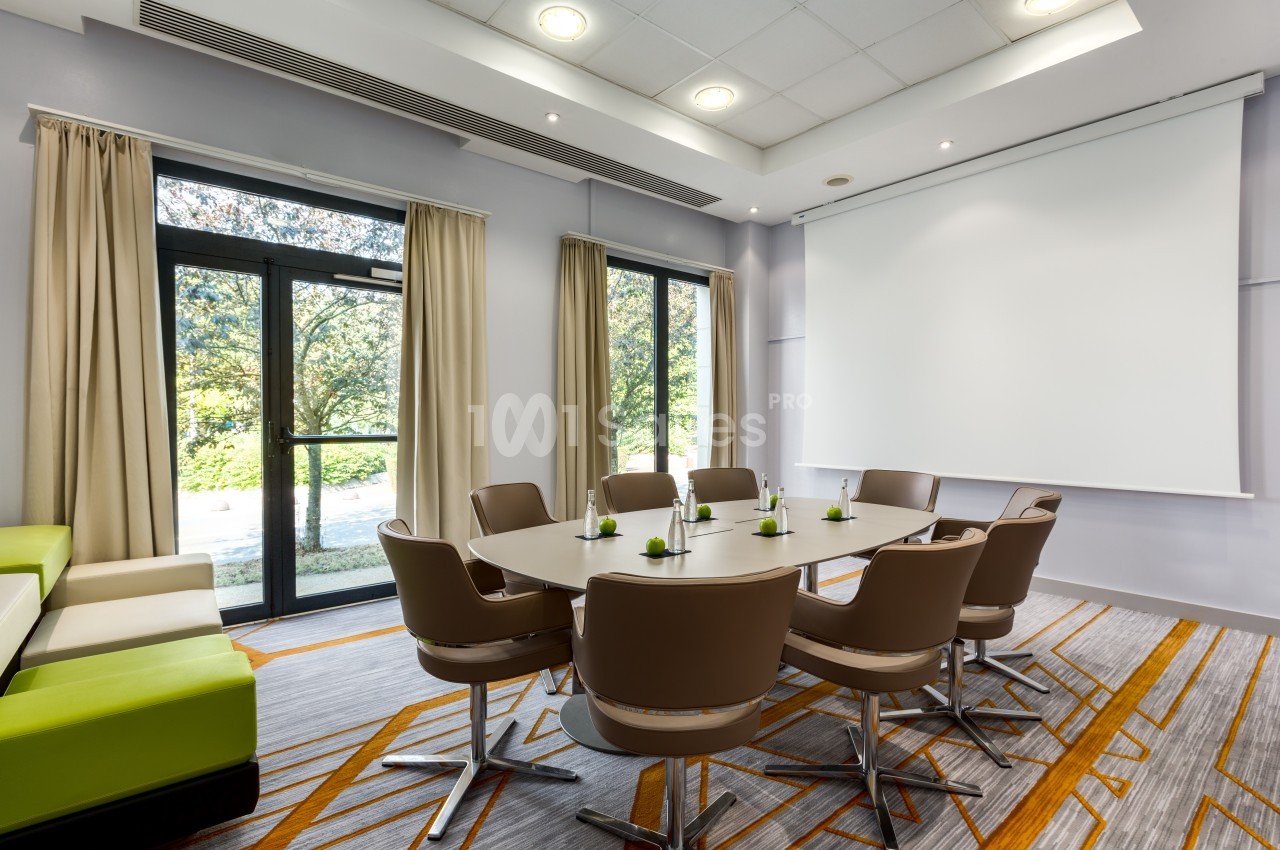 Salle de réunion lumineuse avec table ovale, chaises modernes, rideaux beiges et écran de projection.