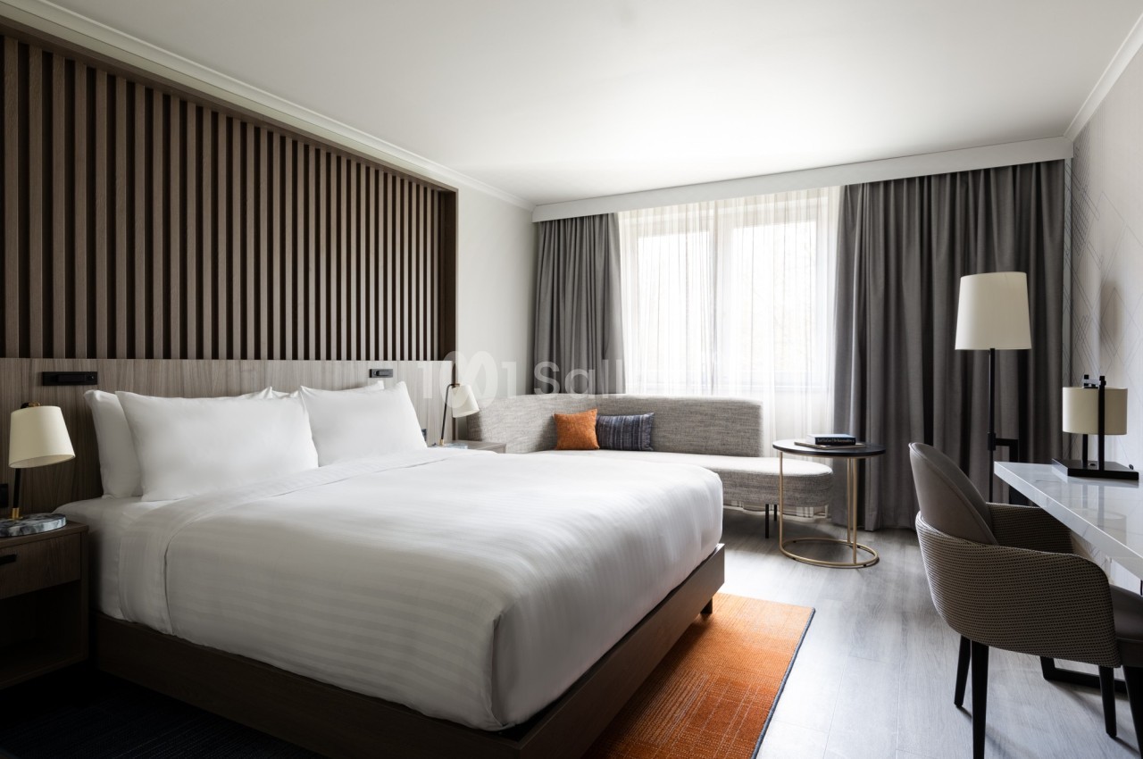 Chambre d'hôtel moderne avec lit double, bureau, canapé, table basse et grande fenêtre avec rideaux.