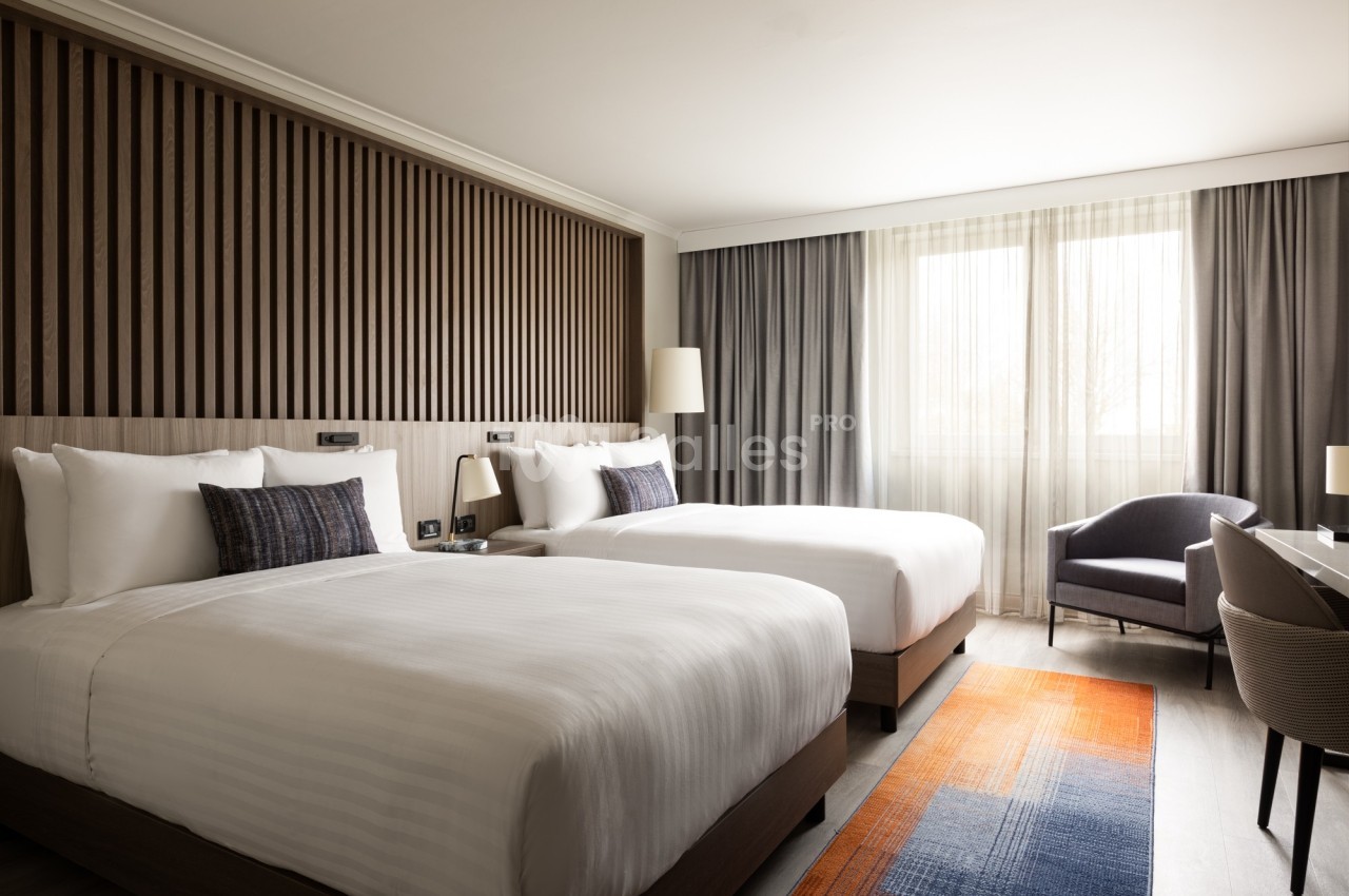 Chambre d'hôtel moderne avec deux lits doubles, un fauteuil, un bureau et une grande fenêtre laissant entrer la lumière…