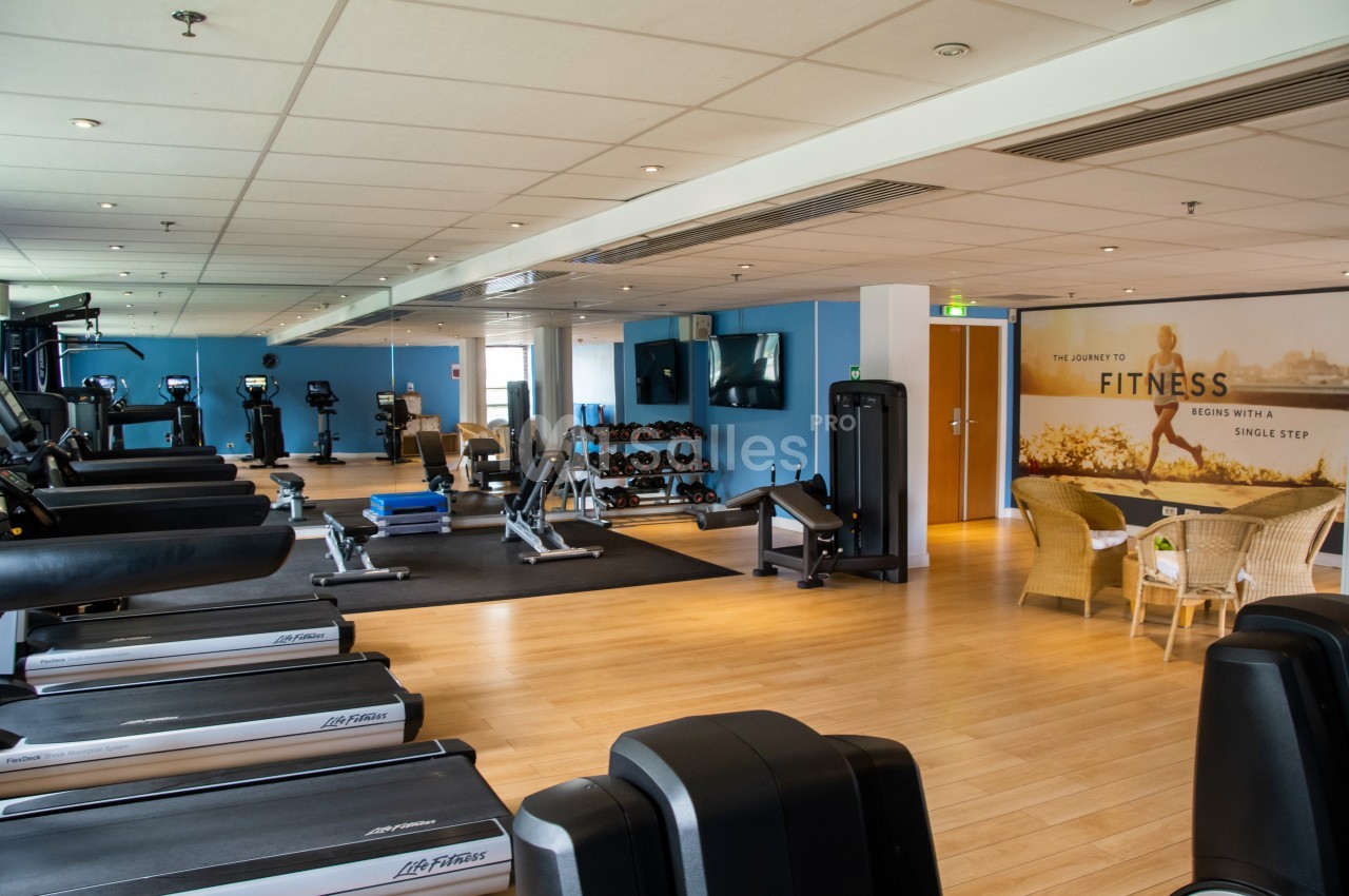 Salle de sport lumineuse avec tapis de course, vélos, haltères et coin détente avec fauteuils en osier.