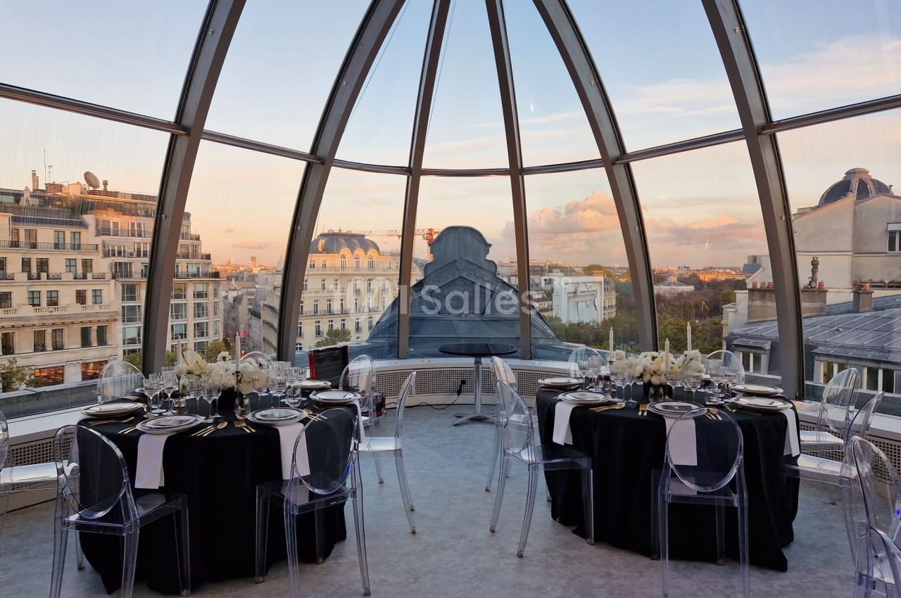 Salle à manger élégante sous une verrière offrant une vue panoramique sur des bâtiments parisiens au coucher du soleil.