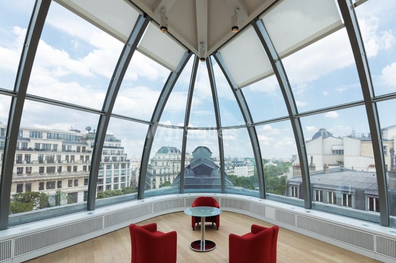 Salle lumineuse avec verrière arrondie, deux fauteuils rouges et une table en verre, vue sur des bâtiments urbains.
