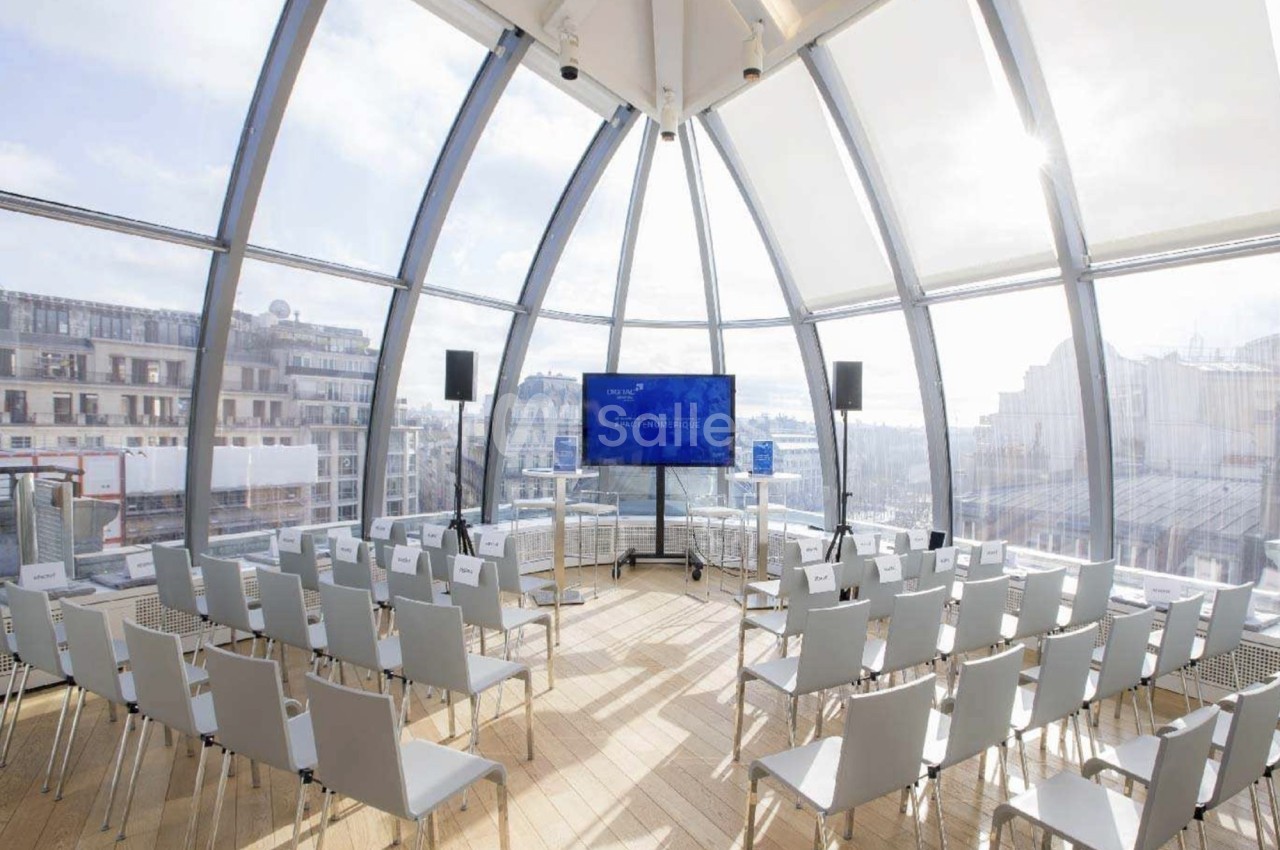 Salle lumineuse avec baies vitrées, équipée de chaises blanches alignées et d'un écran pour une présentation.