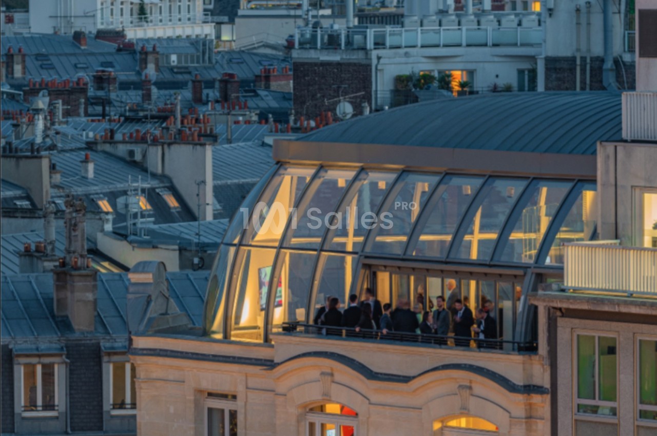 Vue d'une terrasse vitrée éclairée en soirée, surplombant des toits urbains avec des cheminées.