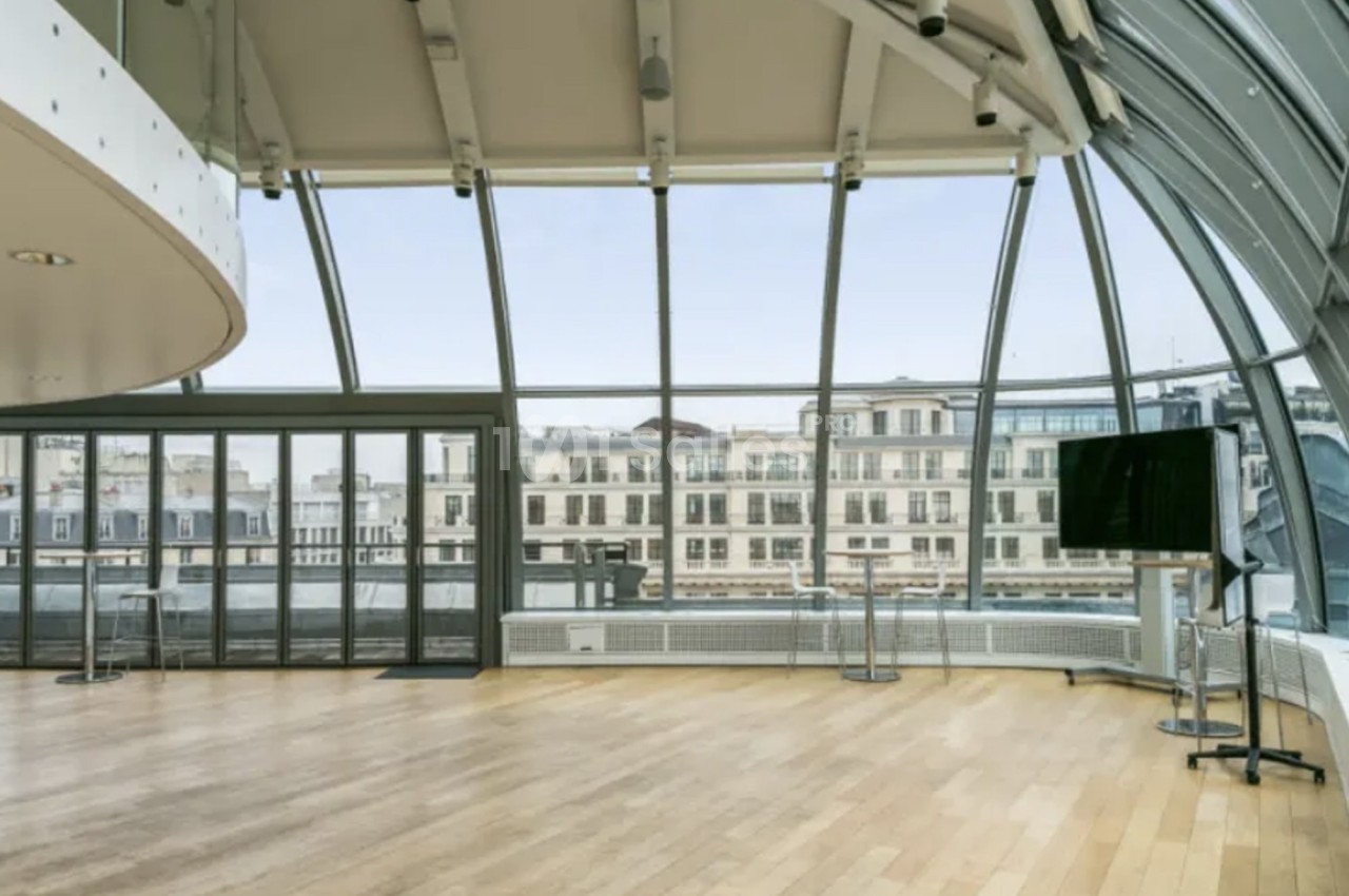 Salle lumineuse avec parquet en bois, grandes baies vitrées courbées et vue sur des bâtiments urbains.
