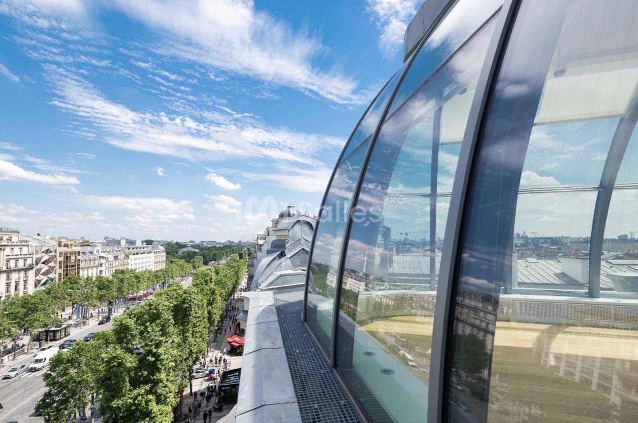 Vue d'une avenue bordée d'arbres et de bâtiments, reflétée dans une façade vitrée sous un ciel dégagé.