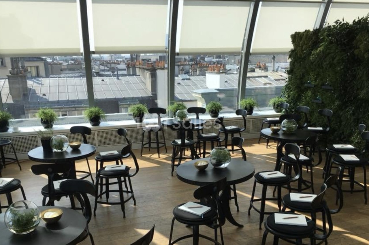 Salle lumineuse avec des tables rondes noires, des chaises assorties, des plantes et une grande baie vitrée donnant sur des…