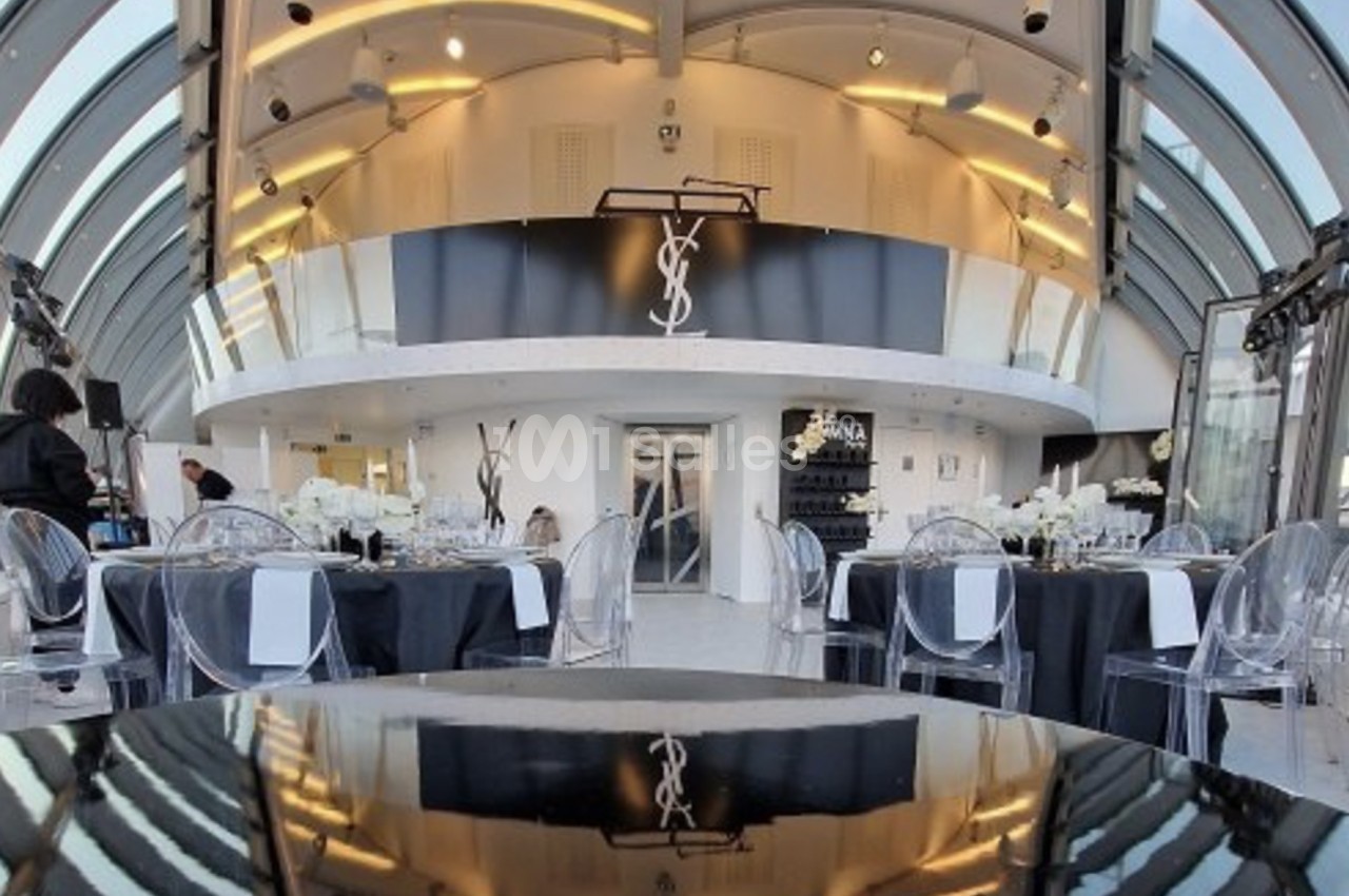 Salle élégante avec tables dressées, chaises transparentes et décoration moderne, dominée par un logo ’YSL’ en hauteur.