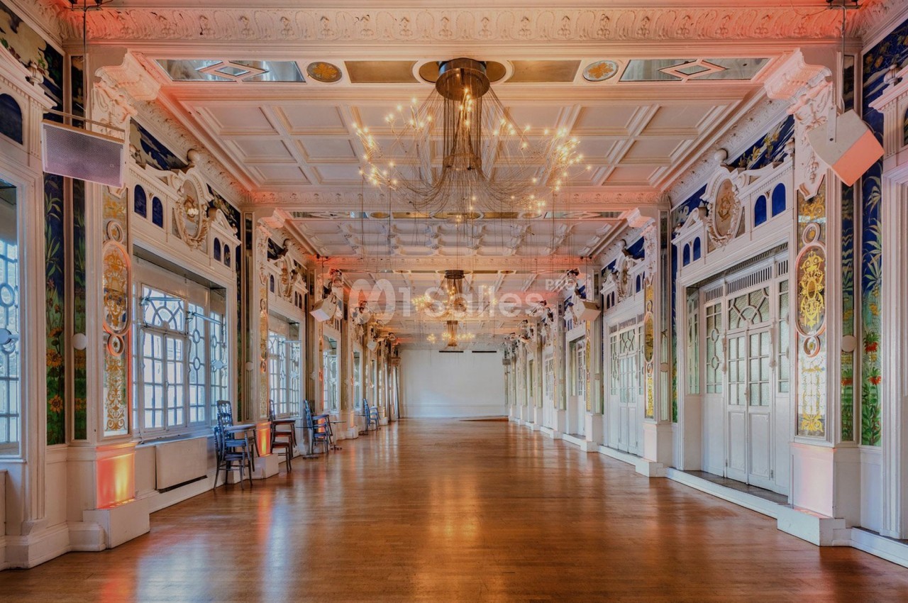 Salle spacieuse avec parquet, lustres suspendus et murs ornés de fresques et de grandes fenêtres.