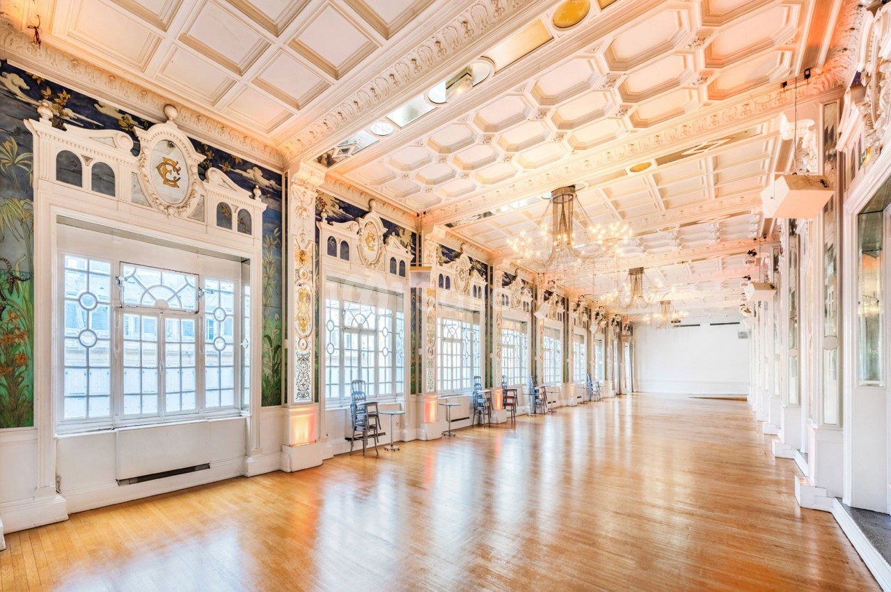 Salle lumineuse avec parquet en bois, grandes fenêtres ornées de vitraux et plafond décoratif avec moulures.
