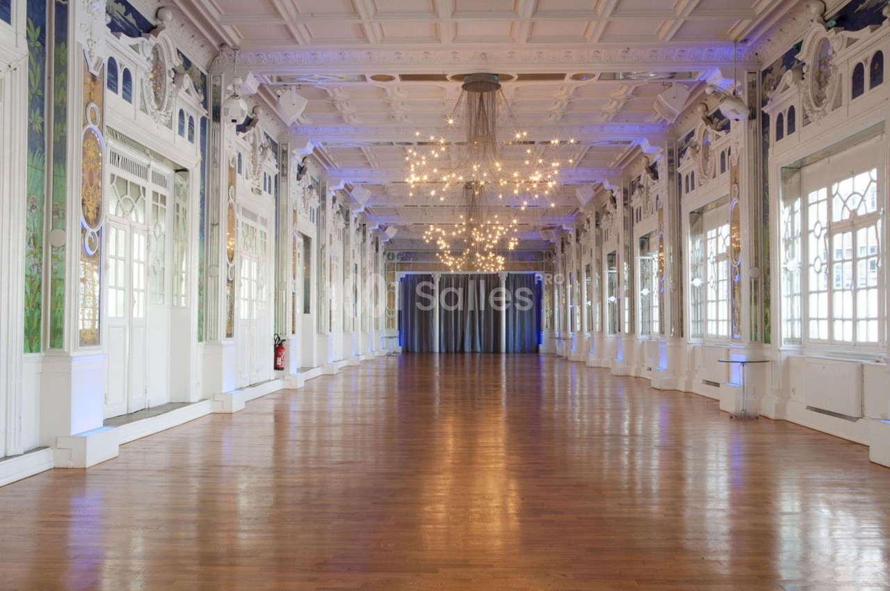 Grande salle lumineuse avec parquet en bois, murs ornés de moulures et lustres suspendus au plafond.