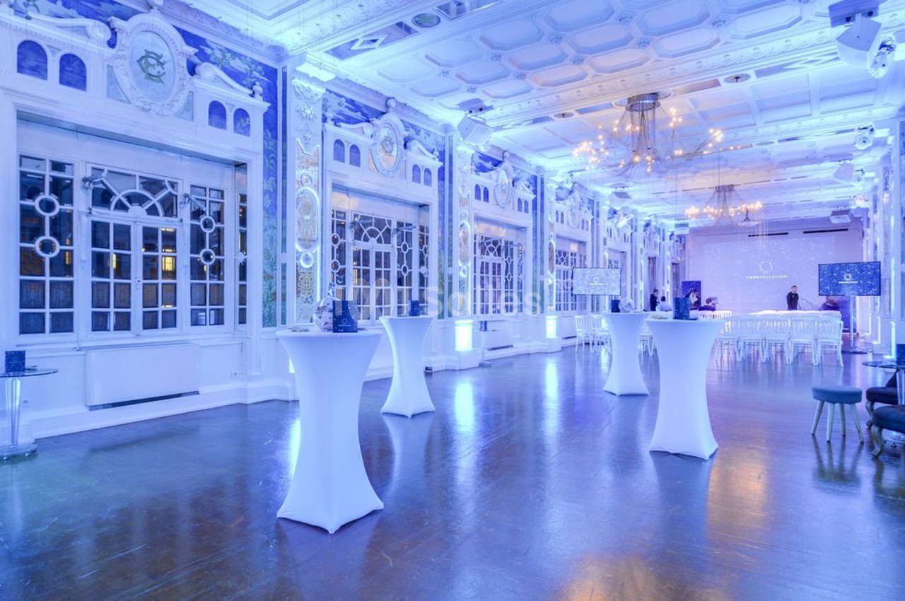 Salle de réception lumineuse avec hauts plafonds, grandes fenêtres, tables hautes et éclairage bleu.
