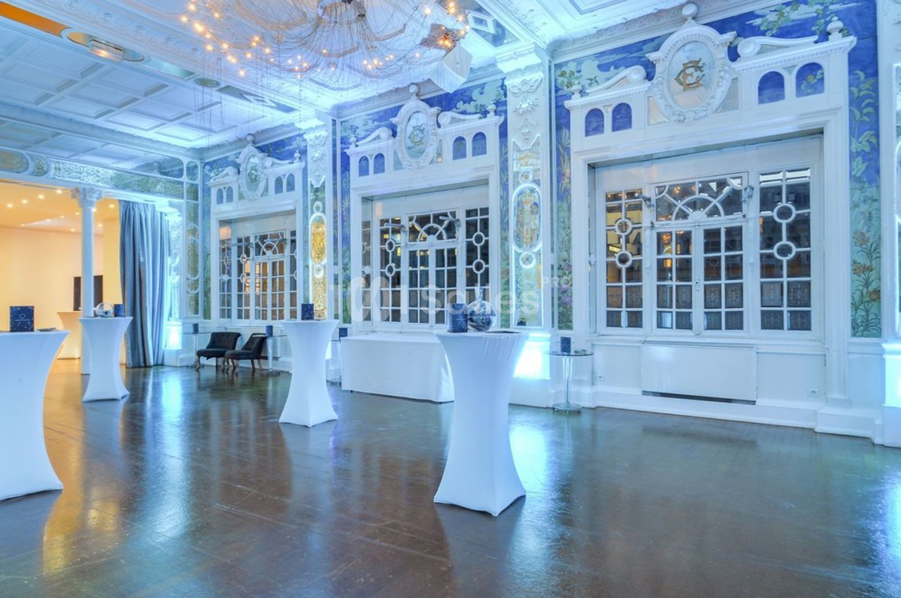 Salle élégante avec hauts plafonds, fresques murales détaillées, grandes fenêtres et tables hautes blanches.