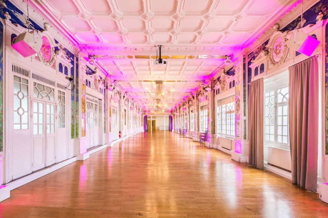 Salle spacieuse avec parquet, murs ornés et éclairage rose, grandes fenêtres laissant entrer la lumière naturelle.