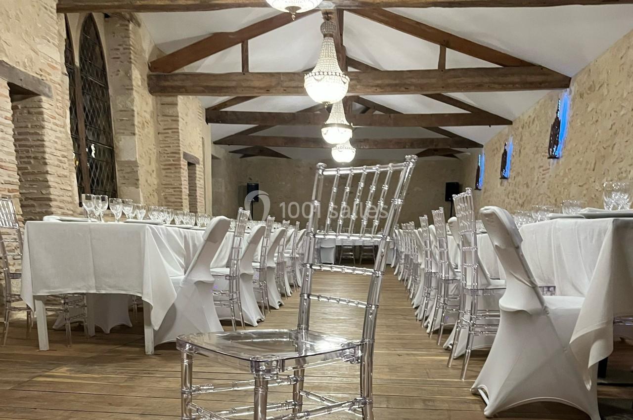 Salle de réception avec tables dressées, nappes blanches et chaises transparentes, éclairée par des lustres suspendus.