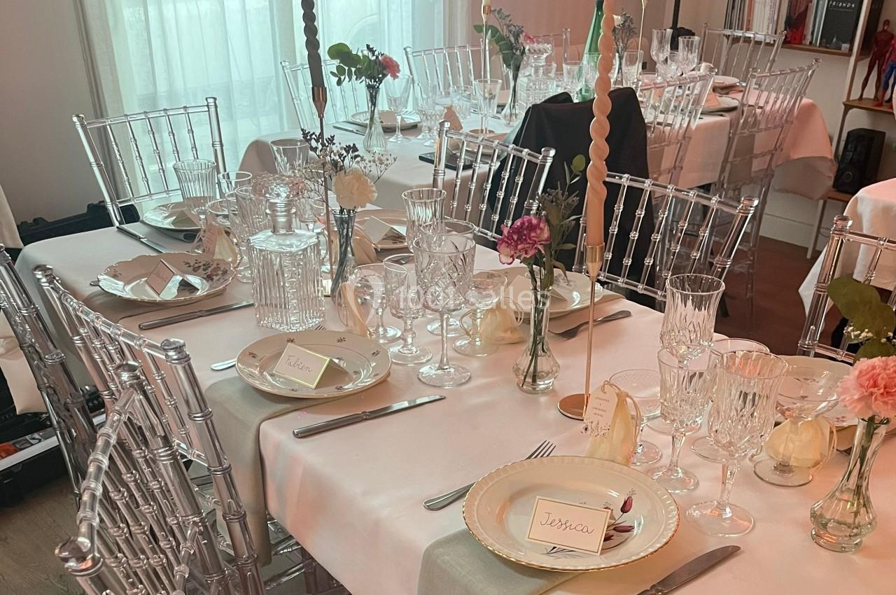 Table élégamment dressée avec nappes blanches, chaises transparentes, vaisselle raffinée, fleurs et bougies décoratives.
